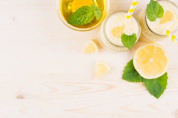 Freshly blended yellow lemon smoothie in glass jars with straw, mint leaf, cut lemon, honey, top view. White wooden board background, copy space.