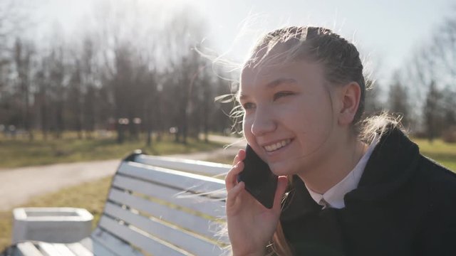 Gimbal shot of female teen girl talking on the phone sitting on bench in spring sunny day, uhd prores footage