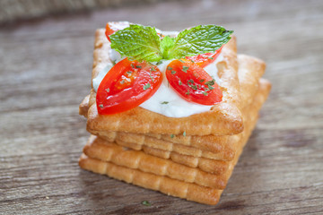 Cracker topped with cream cheese, tomato and mint on wooden background