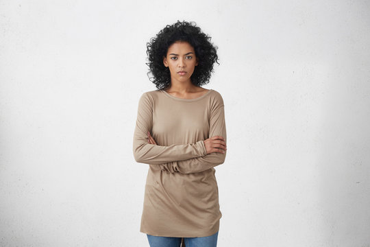 Indoor studio shot of angry young dark-skinned housewife in casual clothes standing in closed posture, keeping arms folded, mad at her drunk husband. Unhappy woman feeling insulted or offended