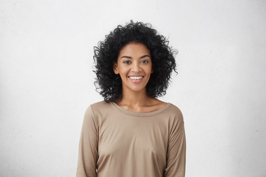 Waist up portrait of cheerful young mixed race female with curly hair posing in studio with happy smile. Dark-skinned woman dressed casually smiling joyfully, showing her white straight teeth