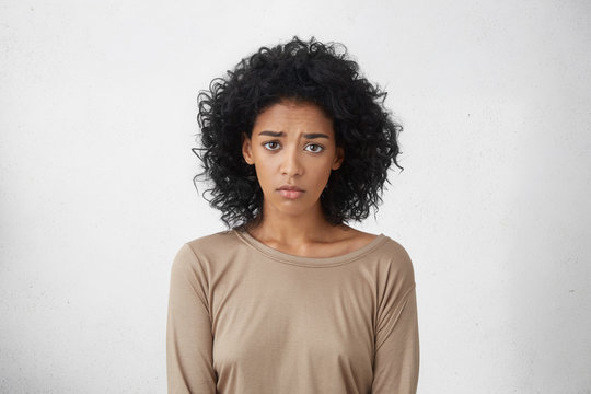 Sad pretty girl feeling upset while spending time at home alone. Beautiful young dark-skinned female with Afro hairstyle staring at camera with unhappy or regretful look against studio wall