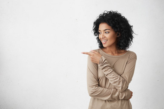Attractive smiling young female customer with curly hair looking sideways and pointing her index finger at copy space on white blank studio wall for your text or promotional content. Horizontal