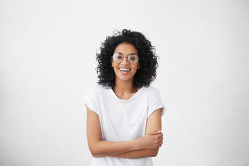 Indoor portrait of beautiful young dark-skinned woman wearing big round eyeglasses and casual white...