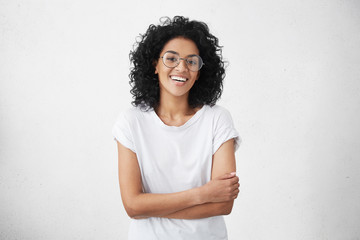 Charming young dark-skinned woman with curly hairstyle having shy smile posing in studio in closed...