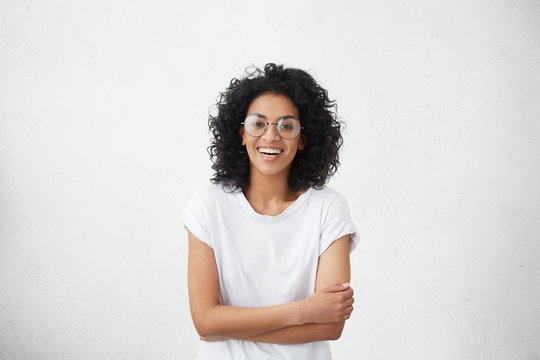 Indoor portrait of beautiful young dark-skinned woman wearing big round eyeglasses and casual white t-shirt smiling joyfully, keeping arms crossed, laughing out loud while watching comedy on TV