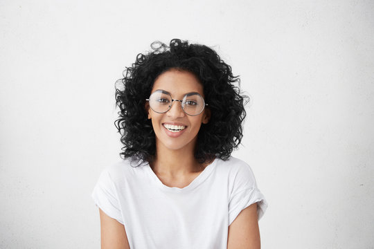 Close up isolated studio shot of good looking cheerful positive young mixed race female with brunette curly hair, smiling broadly, showing her white teeth at camera while having fun indoors