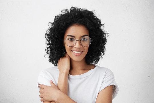Headshot of attractive young mixed race woman wearing big spectacles and white top holding hand behind her neck and looking at camera with subtle smile during awkward conversation with some guy