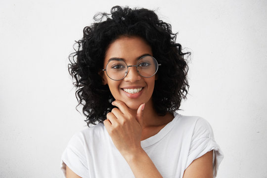 People and lifestyle. Youth and happiness. Close up portrait of confident dark-skinned girl with curly hairstyle posing against white studio wall with copy space for your advertising information