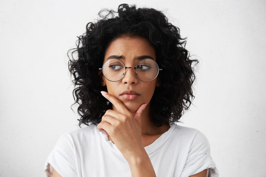 Education, people and life goals. Headshot of attractive mixed race schoolgirl wearing stylish round eyeglasses having doubtful and indecisive look, holding her chin as she has to choose college