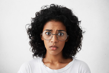 Fototapeta premium Human emotions, feelings, reaction and attitude. Startled mixed race girl wearing big round glasses looking at camera with shocked expression, afraid of sudden noise while staying home alone
