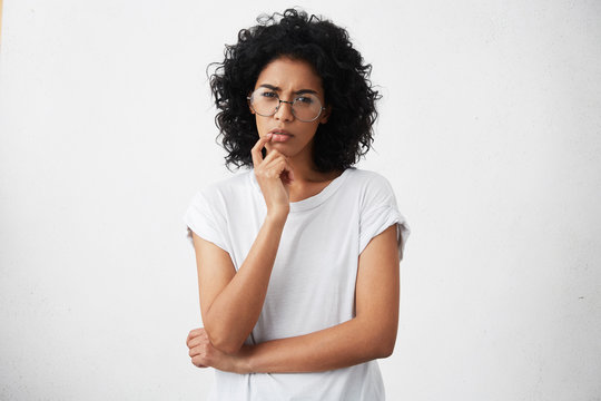 Human emotions, feelings, reaction and attitude. Attractive girl in casual t-shirt and round glasses squeezing eyes and keeping hand on chin in doubt and suspicion, feeling sceptical about something