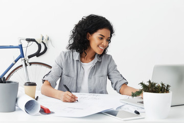 Portrait of positive young African American woman senior architect studying sketches by her...