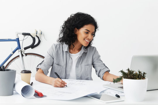 Portrait of positive young African American woman senior architect studying sketches by her trainee, checking measurements on notebook computer and making correction on drawings in front of her