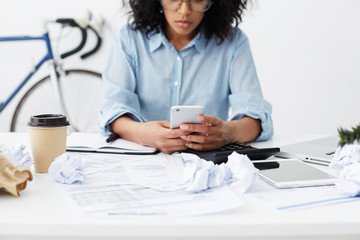 Cropped view of female office worker messaging online, using smart phone while having a rest during lunch break, siting at white office desk with crumpled paper balls and cup of takeaway coffee
