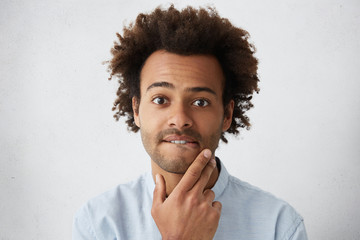 Headshot of handsome troubled or puzzled young African American man with funky hairstyle and stubble biting his lower lip and rubbing chin. Human facial expressions and emotions, body language