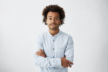 Scared young Afro American man in casual shirt keeping arms folded, biting lips, feeling nervous...