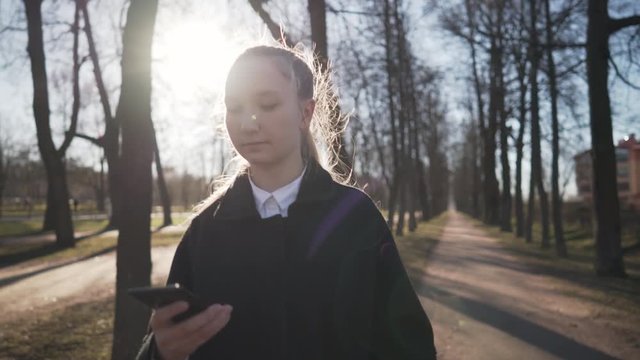 Gimbal shot of female teen girl using smartphone and walking on town alley in spring sunny day, uhd prores footage