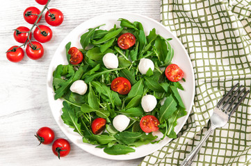 Vegetarian food and healthy lifestyle concept - spring salad with cherry tomato, mozzarella cheese and arugula on a white plate laying on wooden table. Top view, flatlay
