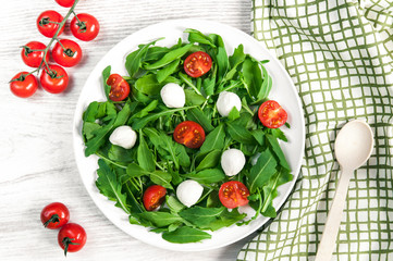 Vegetarian food and healthy lifestyle concept - spring salad with cherry tomato, mozzarella cheese and arugula on a white plate laying on wooden table. Top view, flatlay