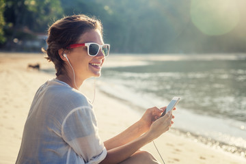 Young beautiful woman hipster traveler in headphones and with mobile phone in hands, on the beach in the sun