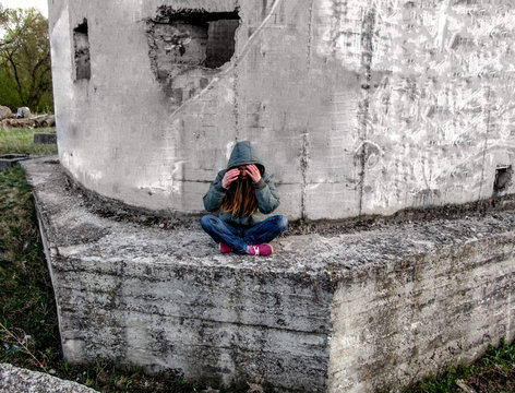 A girl in a hood sits in a lotus pose on a concrete wall. She has long blond hair and she hides her face with her hands