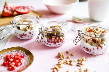 Diet dessert with yogurt, granola and pomegranates in a glassware. Three jars. Horizontal. Rose tablecloth, silver spoon, wooden board. Closeup.