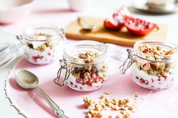 Diet dessert with yogurt, granola and pomegranates in a glassware. Three jars. Horizontal. Rose tablecloth, silver spoon, wooden board. Closeup.