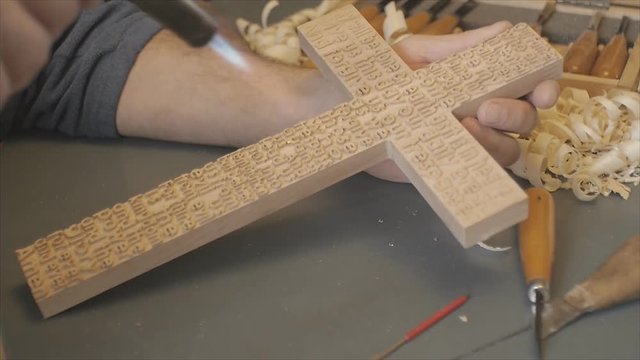 The artist carves a wooden cross with carved prayers are with the tools on dark background.