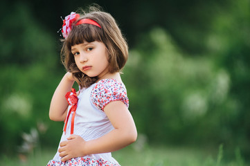 Little girl model in white dress fixes her loose hair with her hand on a nature background.