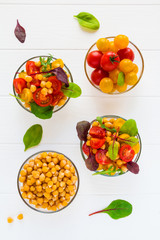 Chickpea salad with fresh red and yellow cherry tomatoes and mix of green salad leaves in glass bowls on the white wooden table. Top view.