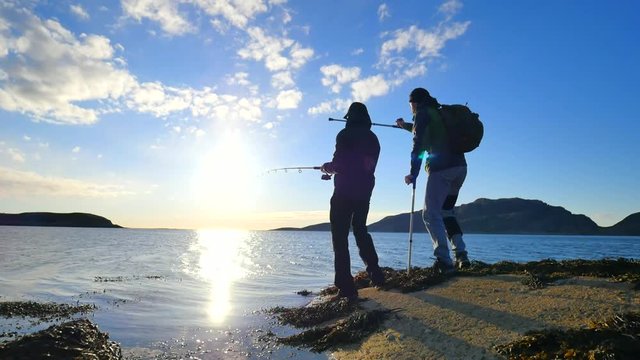 Friends talk about fishing and health. Young working fisherman on stone at sea level. Hiker man with medicine crutches and leg in functional knee braces. 
Men enjoying fishing with beautiful sunset