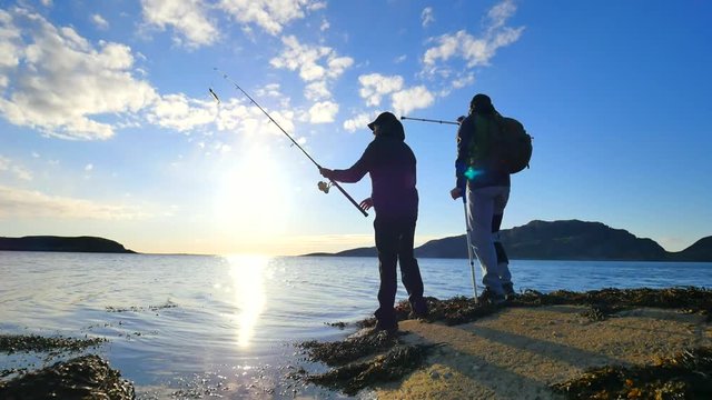 Friends talk about fishing and health. Young working fisherman on stone at sea level. Hiker man with medicine crutches and leg in functional knee braces. 
Men enjoying fishing with beautiful sunset