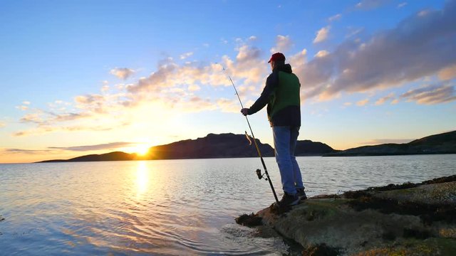 The active man is fishing on sea from the rocky coast. Fisherman check pushing bait on the fishing line, prepare rod and than throw lure into peacefull water. Fisherman silhouette at sunset