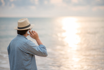 Young Asian man with jean shirt and hat talking on phone and see the sunset on tropical beach,...