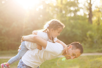 Fototapeta premium Attractive young man is playing with his daughter in the nature. The father is standing and carrying girl on his back. He is stretching arms sideways. The family is smiling