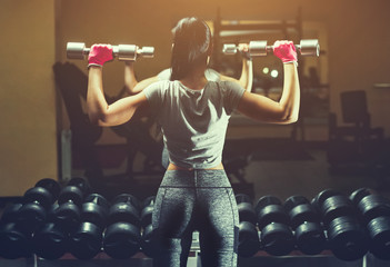 Slim bodybuilder girl lifts heavy dumbbell standing in front of the mirror while training in the...