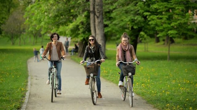 Three Active Young Women are Riding Together in the City Park. Smiling Brunettes with Bicycles Outdoors.