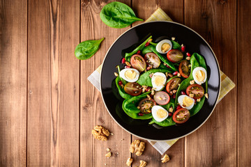 Fresh vegetable salad with spinach, cherry tomatoes, quail eggs, pomegranate seeds and walnuts in black plate on wooden table.
