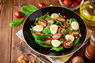 Fresh vegetable salad with spinach, cherry tomatoes, quail eggs, pomegranate seeds and walnuts in black plate on wooden table.