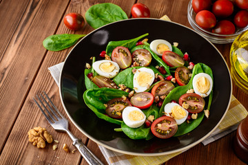 Fresh vegetable salad with spinach, cherry tomatoes, quail eggs, pomegranate seeds and walnuts in black plate on wooden table.