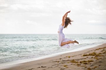 Fototapeta na wymiar Young woman in a white dress jumping on the beach by the ocean. Happy vacation. Beach girl has fun by ocean. Excited girl jumping on white sand by beautiful blue sea.