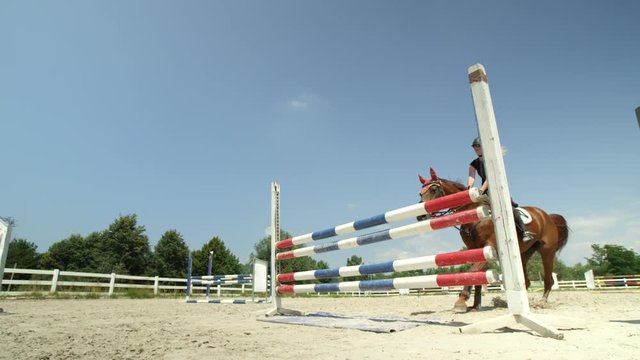 SLOW MOTION, CLOSE UP, LOW ANGLE: Woman riding beautiful chestnut mare jumping the fence in sunny outdoor parkour arena. Horse with a female rider training for the open jumping competition in manege