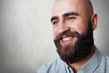 A portrait of handsome bald man with thick beard and mustache having sincere smile while posing against white background. A fashionable bearded hipster looking aside. People and emotions concept.