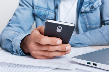 A close-up portrait of male`s hand holding smartphone in his hand dressed in jean shirt sitting in office . A male`s hand with smartphone near business documents and laptop. Business and technology.