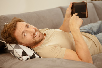Cheerful mature man using a digital tablet lying on the sofa at home