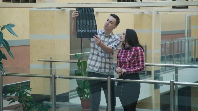 Man explain woman x-ray of different type of sacrum in hall in university. Two students of medical faculty stand on balcony with fence of glass, metal rolls. Tall male with traces of mustache, beard