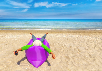 Fototapeta na wymiar Office manager dream about summer beach vacation. Young man dressed sports wear with mobile phone in left hand laying at inflatable mattress on sandy beach in front of beautiful blue sea. Sunny day.