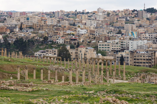 Stone columns and capitals of colonnaded street in the ancient Roman trading city of Jerash, Jordan. The modern city is seen in the background and grassy slopes in the foreground.  Viewed from above.