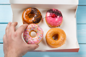 Hand takes donut out of the box with colorful donuts on blue wooden background. Top view